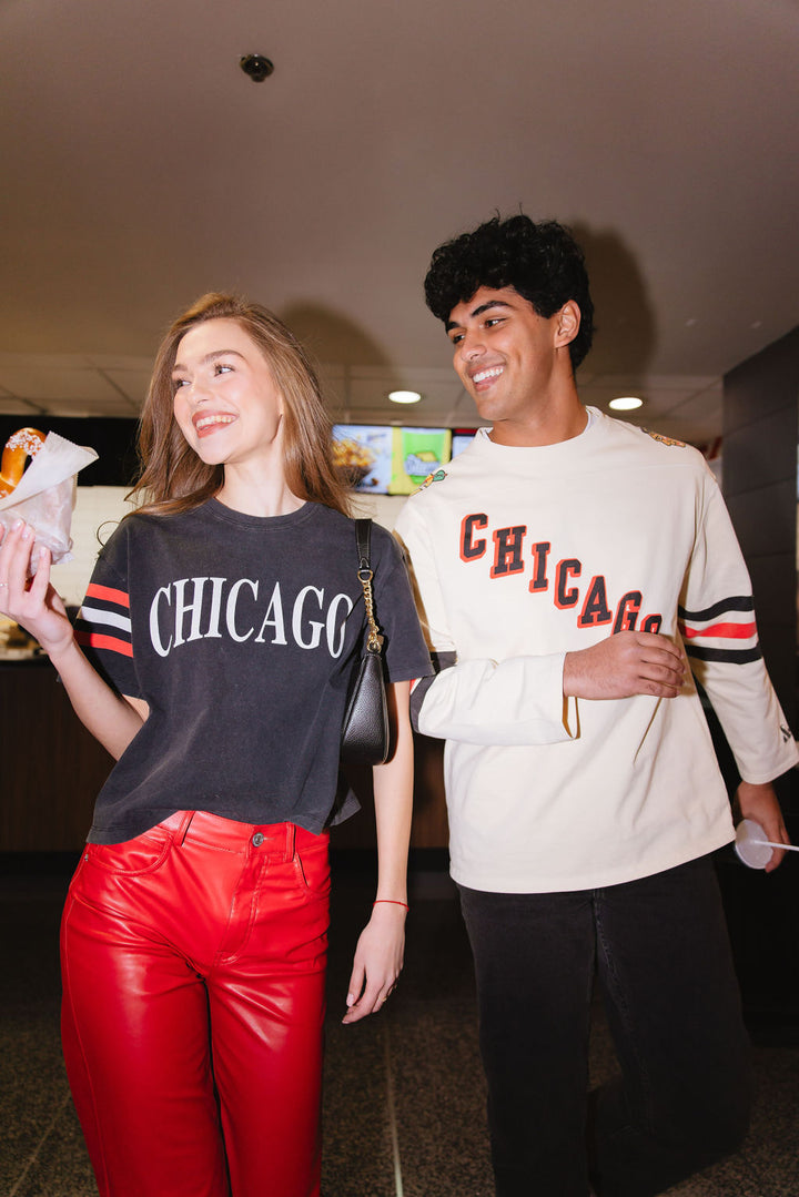 woman wearing black cropped Alice & Wonder tee with Chicago wordmark graphic across front and Chicago Blackhawks primary logo on shoulder with jersey-inspired stripes on right sleeve next to man