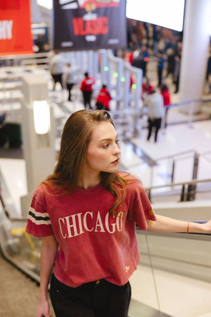 woman wearing red boxy crop Alice & Wonder tee with Chicago wordmark across front and Chicago Blackhawks primary logo on left sleeve and jersey stripes on right sleeve