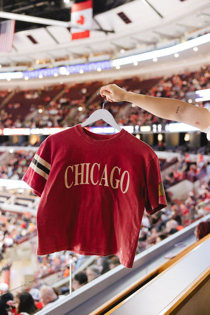 red boxy crop Alice & Wonder tee with Chicago wordmark across front and Chicago Blackhawks primary logo on left sleeve and jersey stripes on right sleeve on hanger in United Center