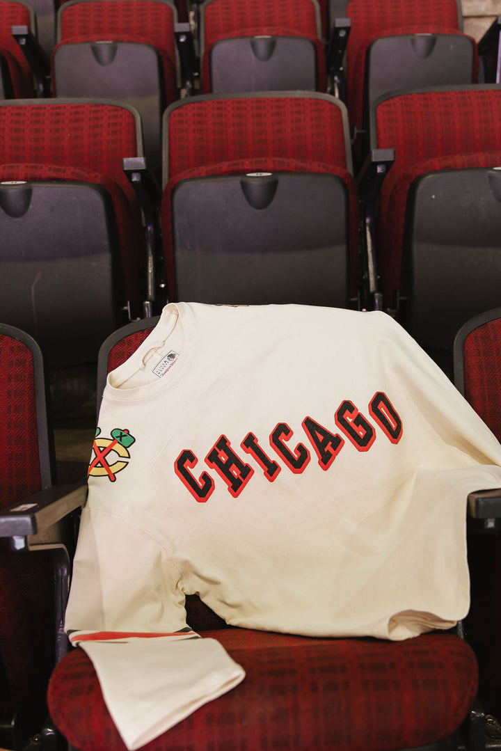 cream Alice & Wonder long sleeve tee with diagonal Chicago wordmark graphic and Chicago Blackhawks secondary and primary logo on shoulders with jersey stripes on sleeves - on seats