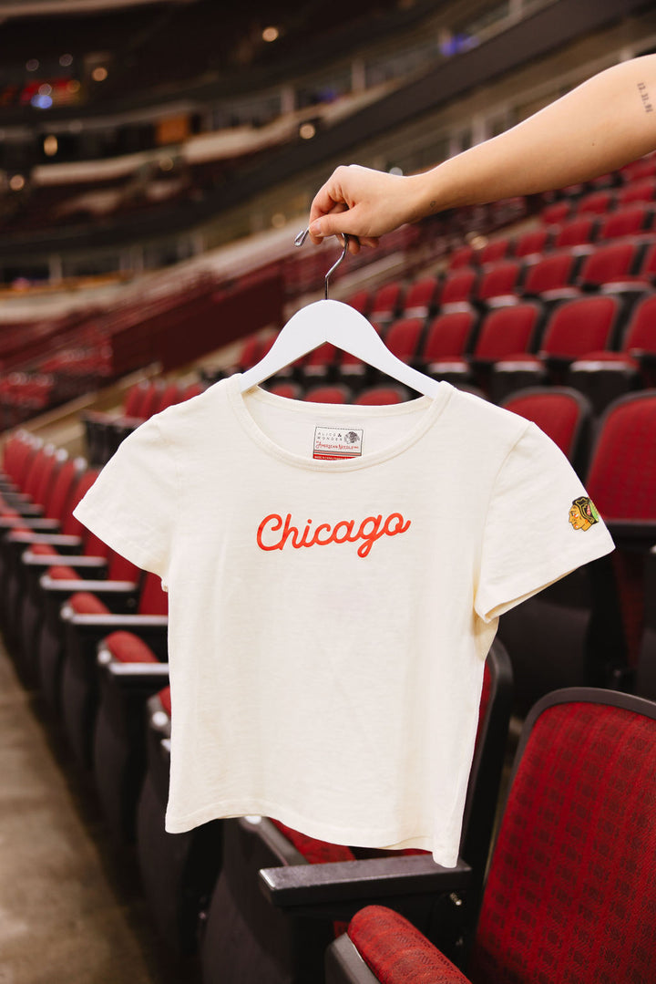 cream Alice & Wonder baby tee with red Chicago script graphic across front and Chicago Blackhawks logo on left sleeve on hanger in front of United Center seats