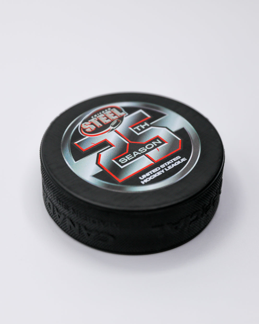 black hockey puck with Chicago Steel logo and 25th season graphic on front - front angled lay flat