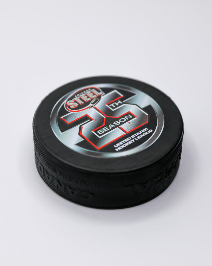 black hockey puck with Chicago Steel logo and 25th season graphic on front - front angled lay flat