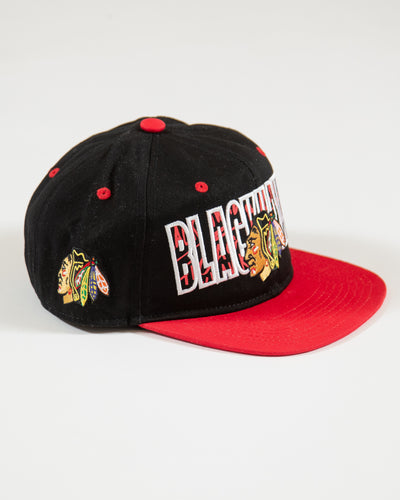Outerstuff black and red youth snapback with flat brim with Chicago Blackhawks wordmark and primary logo with graffiti inspired design - right angle lay flat