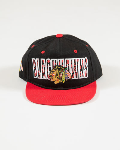 Outerstuff black and red youth snapback with flat brim with Chicago Blackhawks wordmark and primary logo with graffiti inspired design - front angle lay flat