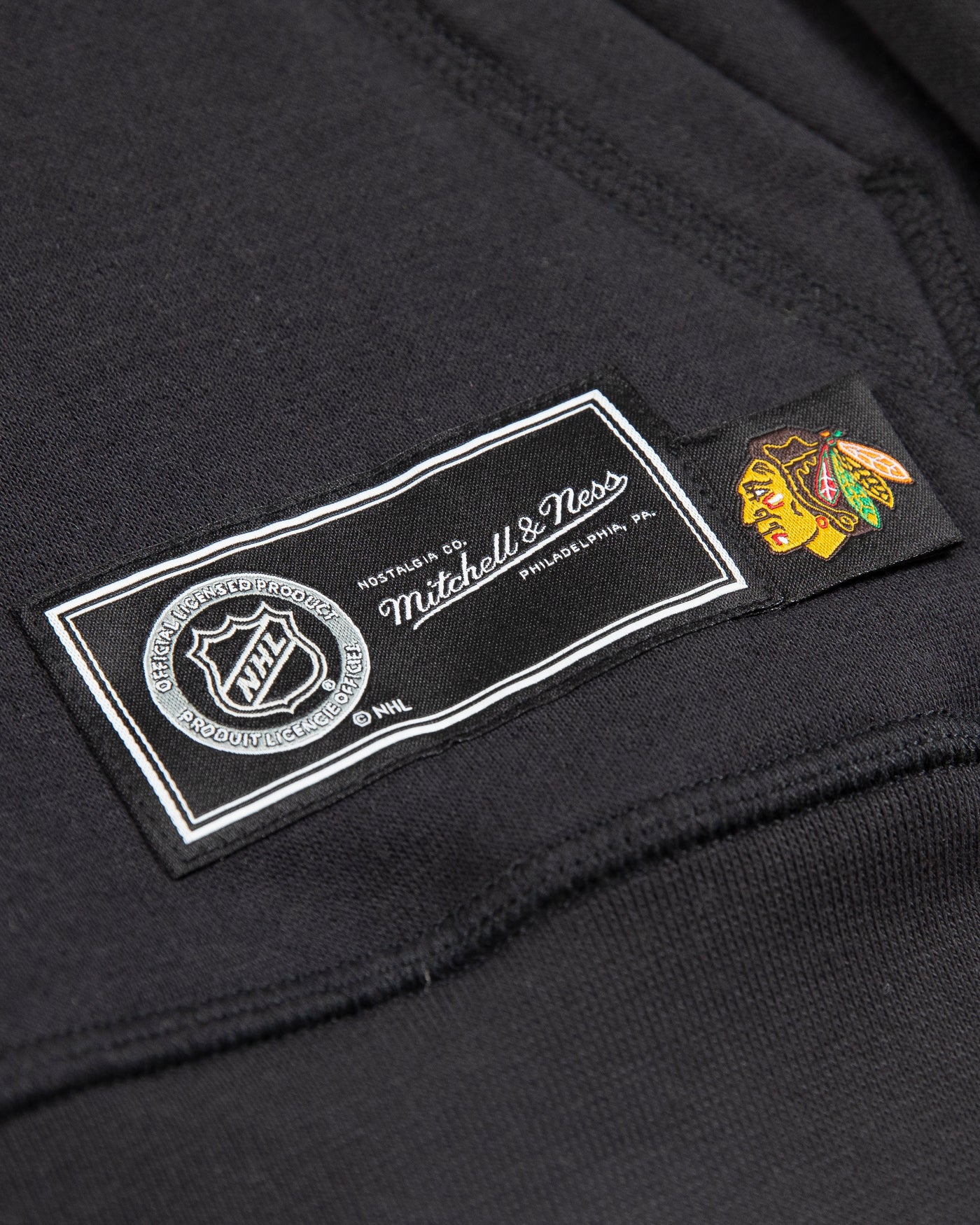 black Mitchell & Ness youth hoodie with Chicago Blackhawks primary logo across chest and Chicago wordmark along left sleeve - NHL logo tag detail lay flat