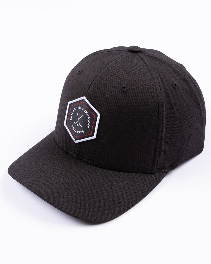 black TravisMathew adjustable cap with hexagon patch on front and Chicago Blackhawks wordmark and crossed hockey sticks graphic - left angle lay flat