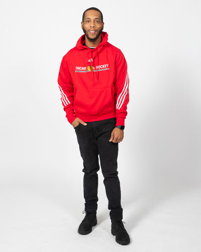 Red adidas hoodie with primary logo and wordmark graphic with three white stripes down sleeves - on model front view