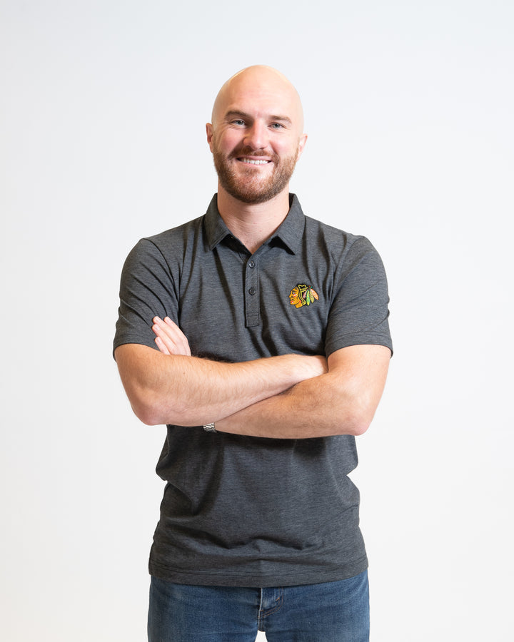 dark grey TravisMathew polo with button closure and embroidered Chicago Blackhawks primary logo on left chest - on model